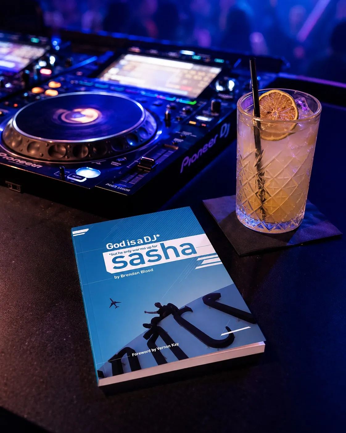 The paperback of God is a DJ* propped against a Pioneer DJ mixer under blue club lighting.